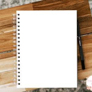 Pode incluir: Um caderno branco com espiral e uma caneta preta sobre uma superf&iacute;cie de madeira. O caderno est&aacute; em branco e pronto para ser escrito.
