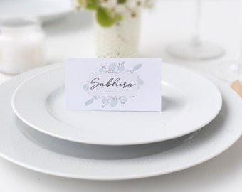 Op de afbeelding: Een witte tafelsetting met een naamkaartje met de tekst "Sabhisa" in script. De kaart is versierd met een bloemenontwerp. Een gouden vork en lepel liggen op tafel. De setting is voor een formeel evenement.