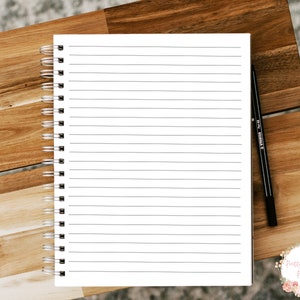 Pode incluir: Um caderno branco de espiral com p&aacute;ginas pautadas e uma caneta preta sobre uma superf&iacute;cie de madeira.