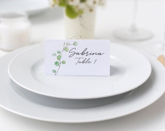 Op de afbeelding: Witte plaatskaart met een groen aquarel bladontwerp. De kaart zegt "Sabrina Tafel 1" in zwarte cursieve letters.