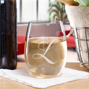 Peut inclure: Un verre à vin sans pied rempli d'un liquide clair, peut-être du vin, avec la lettre "L" cursive blanche gravée sur le devant. Le verre repose sur un tissu blanc, avec une bouteille sombre et un panier en arrière-plan.