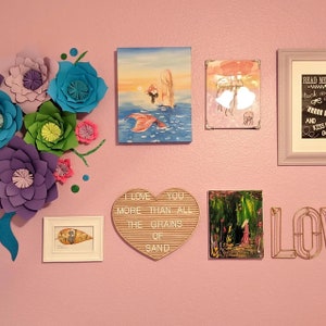 May include: A pink wall decorated with various wall art, including a mermaid painting, a heart-shaped letter board with the text "I love you more than all the grains of sand", a framed print with a jellyfish design, a framed print with the text "Read me tuck me in and kiss goodnight", and a gold metal word "LOVE". The wall is also decorated with paper flowers in various colors, including purple, blue, green, and pink.