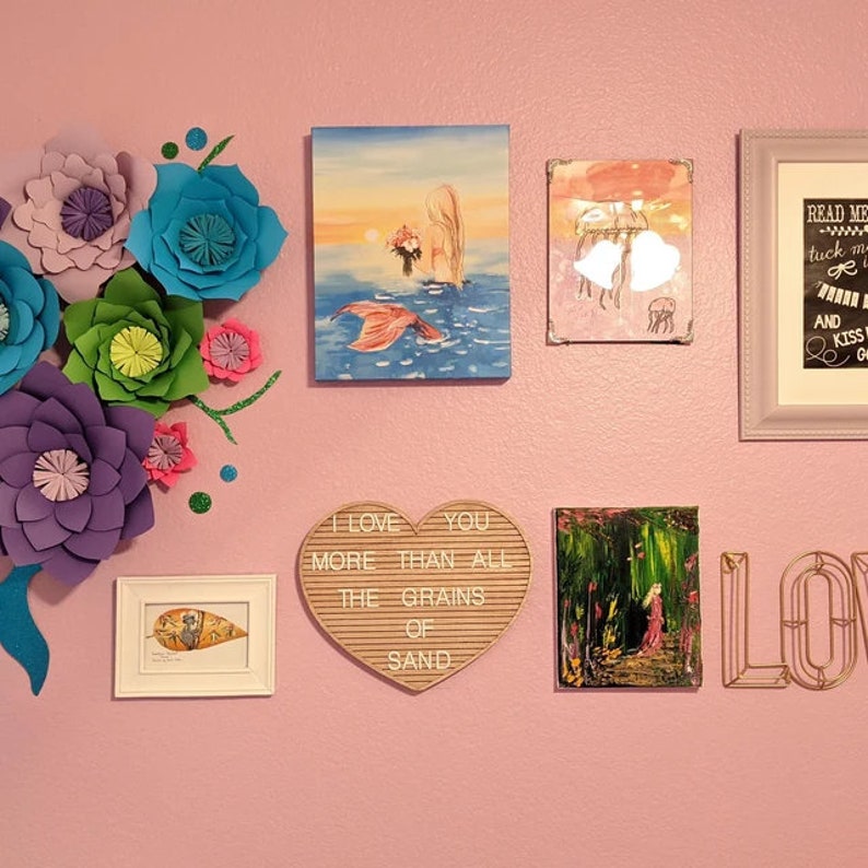 May include: A pink wall decorated with various wall art, including a mermaid painting, a heart-shaped letter board with the text "I love you more than all the grains of sand", a framed print with a jellyfish design, a framed print with the text "Read me tuck me in and kiss goodnight", and a gold metal word "LOVE". The wall is also decorated with paper flowers in various colors, including purple, blue, green, and pink.