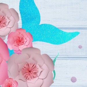 May include: Pink paper flowers with a blue glitter mermaid tail on a light blue wooden background.