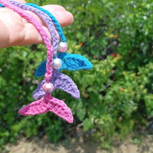 May include: Three crocheted mermaid tails in pink, purple, and blue with pearl beads. The tails are hanging from a hand.