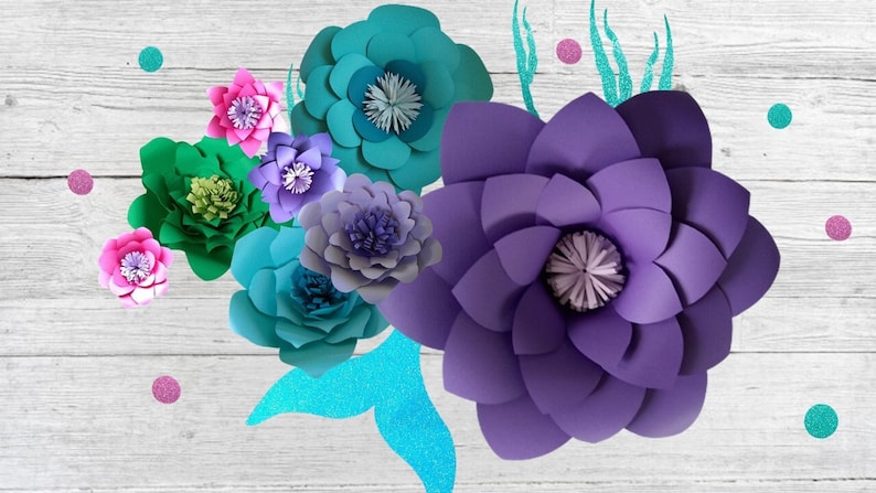 May include: A collection of paper flowers in various colors, including purple, teal, green, and pink. The flowers are arranged on a white wooden background with a teal and silver mermaid tail.
