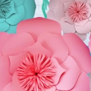 May include: A large pink paper flower with a smaller pink center, surrounded by smaller teal paper flowers. The flowers are against a light blue wooden background.