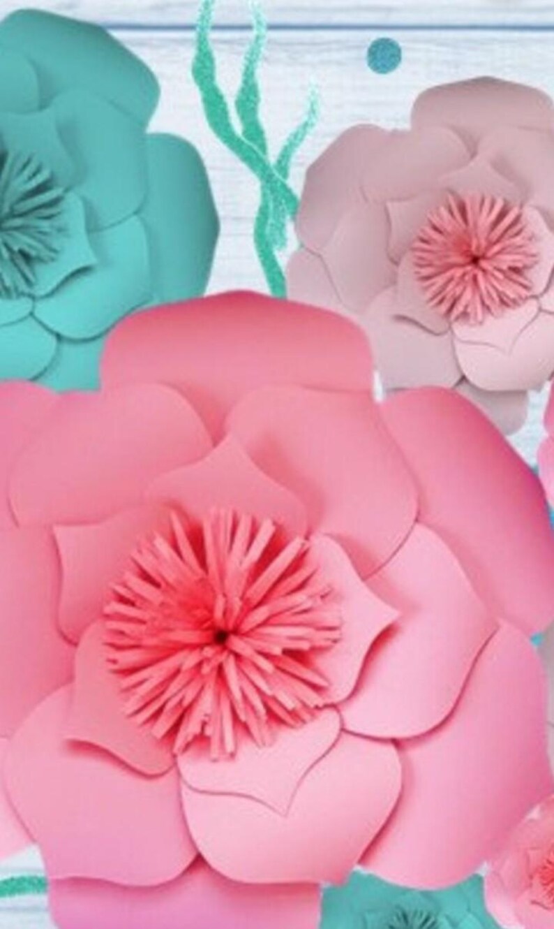 May include: A large pink paper flower with a smaller pink center, surrounded by smaller teal paper flowers. The flowers are against a light blue wooden background.