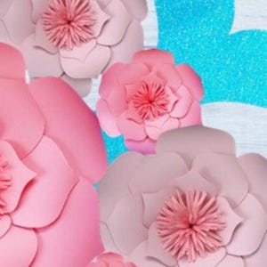 May include: A set of pink and teal paper flowers. The flowers are layered and have a ruffled texture. The flowers are arranged on a blue and white background.