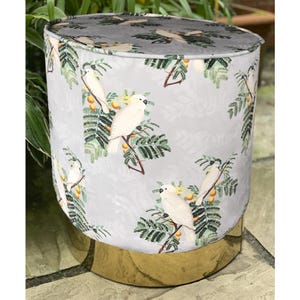 May include: A round ottoman with a light gray fabric featuring a repeating pattern of white birds perched on branches with yellow fruit. The base is gold-colored, and the ottoman is placed on a stone surface.
