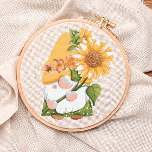 Può includere: Ricamo a punto croce di uno gnomo con un girasole. Lo gnomo indossa un cappello giallo e un abito verde. Il ricamo è posto in un telaio di legno su uno sfondo di tessuto beige.
