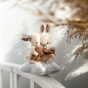 May include: Two plush white bunny rabbits cuddling on a white cloud-shaped pillow. The bunnies have brown ears and noses. The bunny on the left is wearing a brown felt flower.