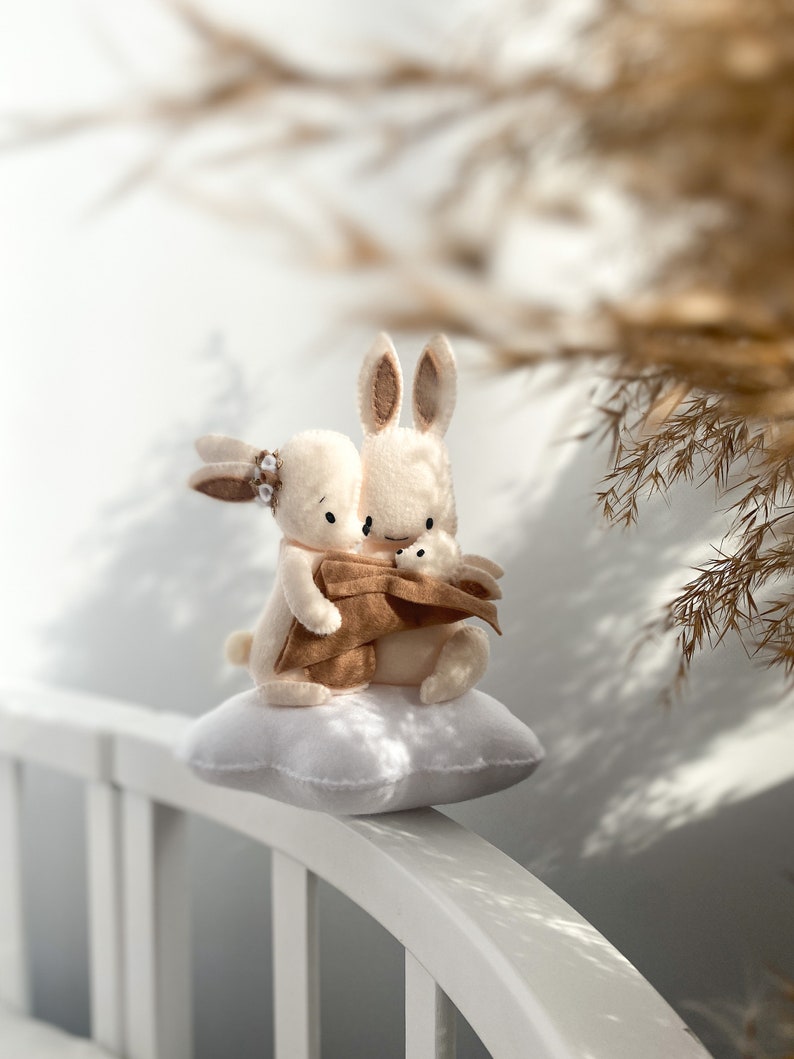 May include: Two plush white bunny rabbits cuddling on a white cloud-shaped pillow. The bunnies have brown ears and noses. The bunny on the left is wearing a brown felt flower.