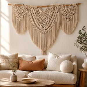 Op de afbeelding: Een grote, crèmekleurige macramé wandhanger opgehangen aan een natuurlijke houten tak. Het stuk bevat ingewikkelde knooppatronen, waaronder geometrische vormen en lange, vloeiende franjes die in drie afzonderlijke secties naar beneden hangen.