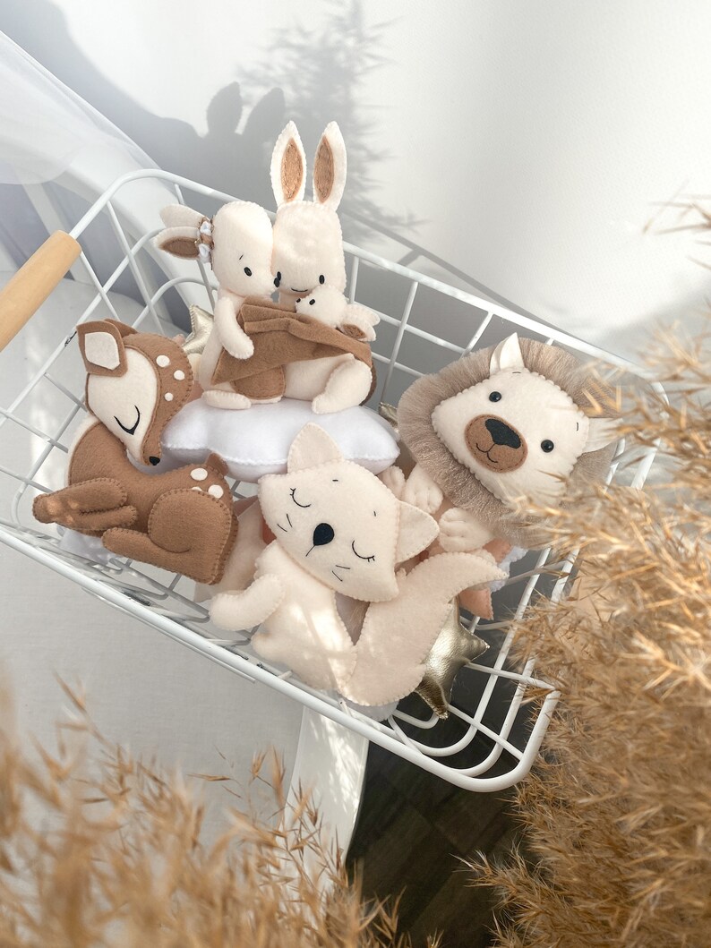 May include: A white wire basket filled with plush animal toys. The toys include a brown deer, a white cat, a brown rabbit, a white rabbit, and a brown and white lion. The toys are all lying down and have embroidered eyes and noses.