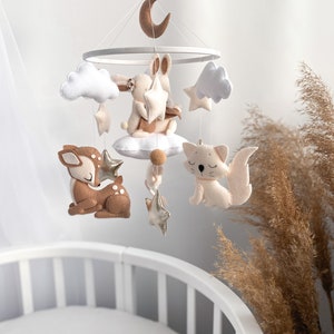 May include: A white and brown felt baby mobile with a crescent moon, clouds, stars, a bunny, a deer, and a cat. The mobile is hanging above a white crib.