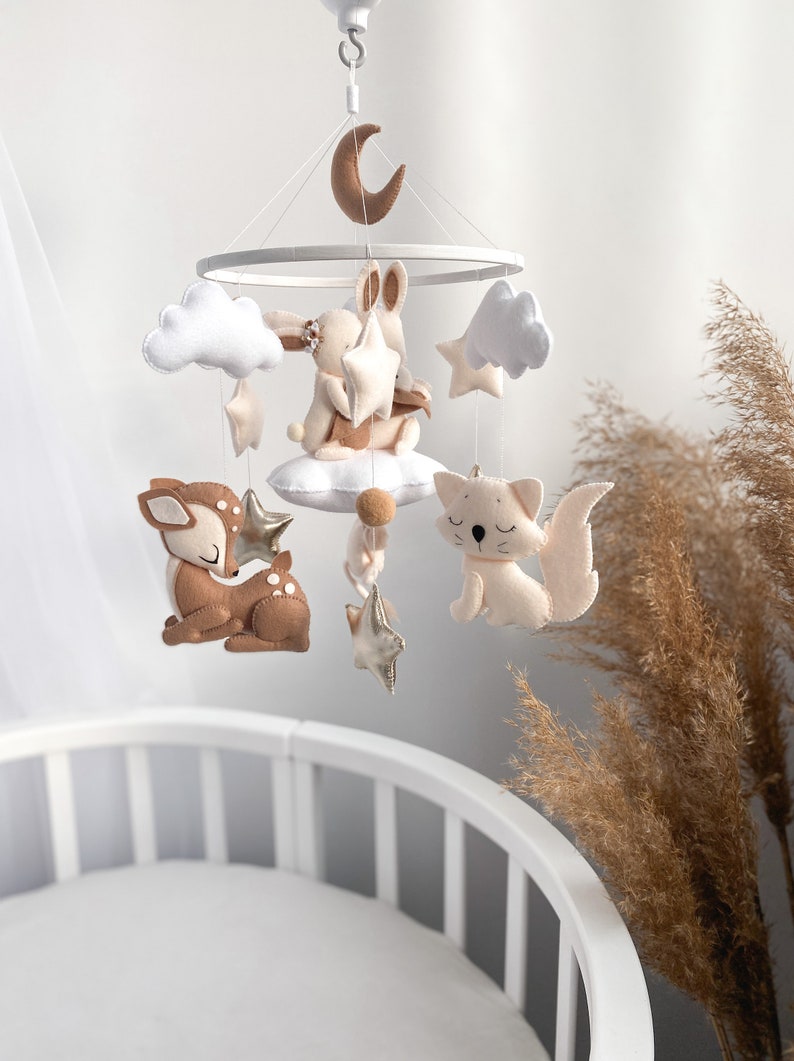 May include: A white and brown felt baby mobile with a crescent moon, clouds, stars, a bunny, a deer, and a cat. The mobile is hanging above a white crib.