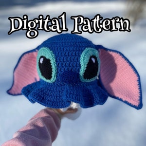 May include: Blue and pink crocheted hat with a cartoon face, resembling a popular animated character. The hat has large ears and a wide brim.