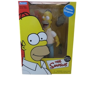 May include: A boxed Homer Simpson action figure from the Faces of Springfield series. The box is yellow with a large cartoon image of Homer's face. The figure is wearing a white shirt, blue pants, and has a surprised expression.