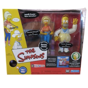 May include: A boxed set of The Simpsons action figures, featuring Beverly D'Angelo and Homer Simpson. The packaging is yellow and red, with the show's logo and character illustrations. The box includes the text "Intelli-Tronic Voice Activation" and "Interactive Mobile Home Environment."