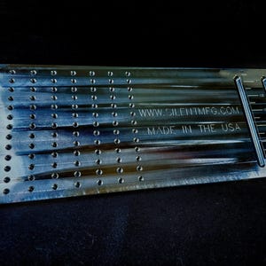 Puede incluir: Una herramienta metálica rectangular con múltiples agujeros perforados y líneas grabadas. La herramienta tiene el texto "WWW.SILENTMFG.COM" y "MADE IN THE USA". La herramienta tiene marcas de medición en el lateral.