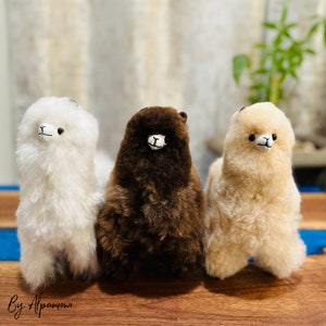 Op de afbeelding: Drie pluche alpaca speelgoed in wit, bruin en beige. Elke alpaca heeft een klein, geborduurd gezicht met zwarte ogen en een lachende mond. De alpaca's staan op een houten oppervlak.