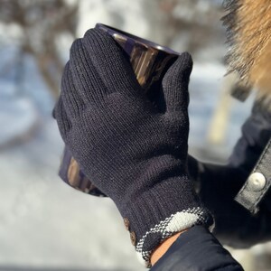 May include: Black knit gloves with a white and black plaid cuff and a button detail. The gloves are being worn by a person holding a mug.