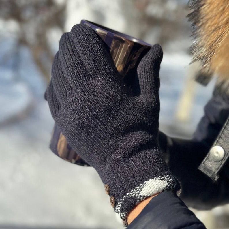 May include: Black knit gloves with a white and black plaid cuff and a button detail. The gloves are being worn by a person holding a mug.
