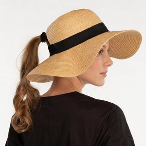 May include: A straw sun hat with a wide brim and a black band. The hat is designed to accommodate a ponytail, with an opening at the back. The hat is a light tan color, and the band is black. The hat is a stylish accessory for sun protection.