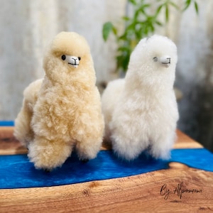 Op de afbeelding: Twee pluche alpaca figuren, een cr&egrave;me en een lichtbruine, staan naast elkaar op een houten oppervlak met een blauwe harsrivier. De alpaca's hebben kleine zwarte ogen en geborduurde monden. De tekst "By Alpamowi" staat rechtsonder.