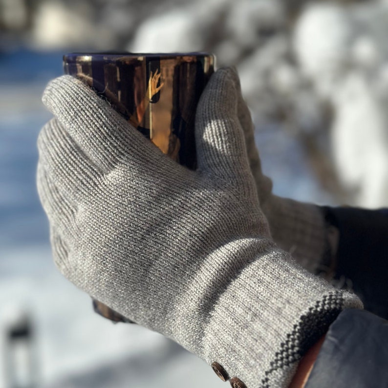 May include: A pair of gray knitted gloves with a black trim. The gloves are being worn by a person holding a mug.