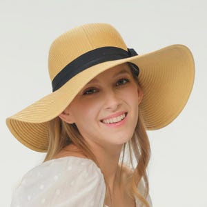 May include: A wide-brimmed, straw-colored sun hat with a black ribbon band. The hat has a rounded crown and a wide, gently curved brim. The black ribbon is tied in a bow on the side. The hat is a stylish accessory.