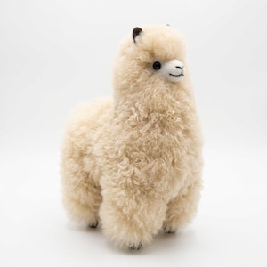 Op de afbeelding: Een cr&egrave;me-kleurig alpaca-knuffel met een pluizige, krullende textuur. De alpaca heeft een wit gezicht, zwarte ogen en kleine zwarte oren. Hij staat op vier korte poten en het algehele ontwerp is zacht en uitnodigend.