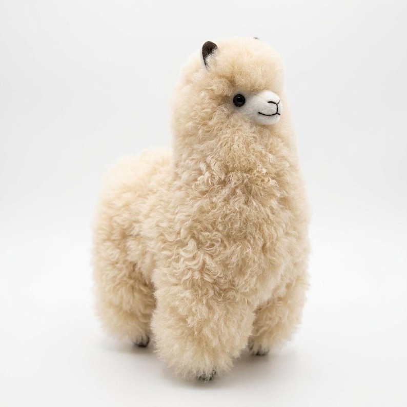 Op de afbeelding: Een cr&egrave;me-kleurig alpaca-knuffel met een pluizige, krullende textuur. De alpaca heeft een wit gezicht, zwarte ogen en kleine zwarte oren. Hij staat op vier korte poten en het algehele ontwerp is zacht en uitnodigend.