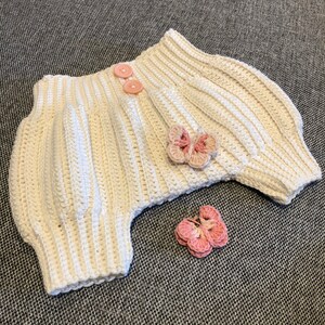 Puede incluir: Unos bloomers de bebé de ganchillo blanco con adornos de flores de ganchillo rosa.