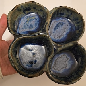 May include: A handmade ceramic serving dish with four connected compartments. The dish has a dark blue glaze with lighter blue and green accents, creating a unique, organic appearance. The dish is ideal for serving snacks or appetizers.