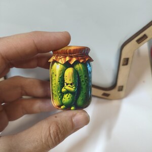 Op de afbeelding: Een kleine, platte, uitgesneden sticker of speld met een cartoonillustratie van een glazen pot gevuld met groene augurken. In de pot staat een enkele, antropomorfe augurk met een norse gezichtsuitdrukking en gekruiste armen centraal.