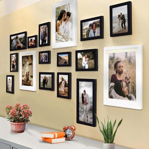 May include: A gallery wall with 14 black and white picture frames of various sizes. The frames are arranged in a staggered pattern on a light beige wall. The frames contain photos of couples, landscapes, and portraits.