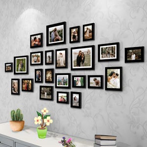 Könnte beinhalten: Eine Bilderwand mit 20 schwarzen Bilderrahmen in verschiedenen Größen, die jeweils ein Foto eines Paares zeigen. Die Rahmen sind in einem versetzten Muster an einer weißen Wand angeordnet. Eine kleine Topfpflanze steht auf einem Regal unter den Rahmen.