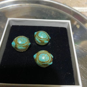 May include: Three turquoise blue turtle-shaped beads in a white box with a black interior. The beads are smooth and have a glossy finish.