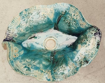 May include: A ceramic sink with a unique, organic shape and a teal glaze. The sink has a decorative design of white flowers and a small drain hole in the center.