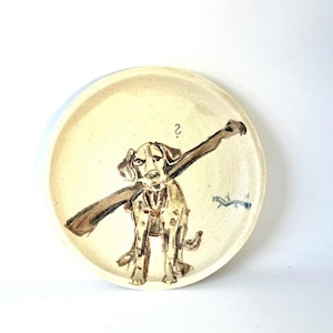 Könnte beinhalten: Keramikplatte mit handgemalter Illustration eines Hundes, der einen Stock trägt. Der Hund ist braun und weiß, und die Platte hat einen cremefarbenen Hintergrund. Eine kleine blaue und braune Illustration eines Fuchses ist ebenfalls vorhanden.