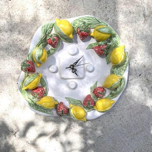 May include: A white ceramic wall clock with a fruit design. The clock face is surrounded by a wreath of yellow lemons, red strawberries, and green leaves. The clock hands are black.