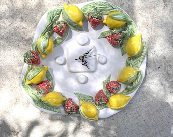 May include: A white ceramic wall clock with a fruit design. The clock face is surrounded by a wreath of yellow lemons, red strawberries, and green leaves. The clock hands are black.