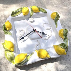 May include: White square ceramic clock with yellow lemon and green leaf accents. The clock face has black hands, a red second hand, and white dotted numbers. The lemons are arranged around the clock's perimeter.