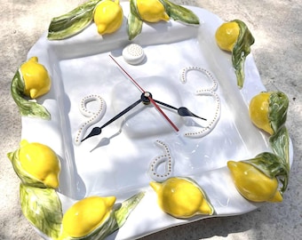 May include: White square ceramic clock with yellow lemon and green leaf accents. The clock face has black hands, a red second hand, and white dotted numbers. The lemons are arranged around the clock's perimeter.