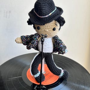 Puede incluir: Muñeca de ganchillo de un artista con sombrero negro, chaqueta con detalles coloridos y pantalón negro con rayas blancas. La muñeca posa con un brazo extendido, sosteniendo un micrófono, sobre un disco de vinilo.