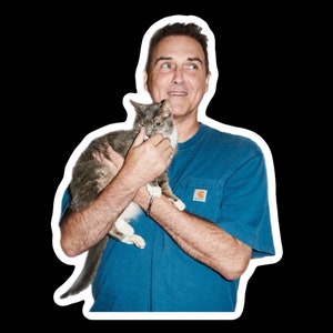 May include: A man wearing a blue t-shirt with a patch on the pocket is holding a gray and white cat.