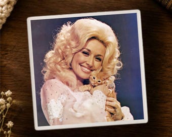 May include: A square coaster featuring a vintage-style photograph of a person with voluminous blonde hair wearing a light pink long-sleeved garment, smiling while holding a small orange kitten against a dark blue background.