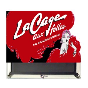 May include: A red and black framed print of the Broadway musical "La Cage aux Folles". The print features a woman in a red feather boa and a pink turban.